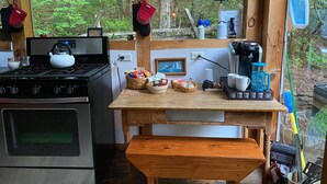 A rustic cabin kitchen with a gas stove, kettle, coffee maker, and various kitchen items on a wooden table.