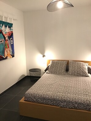 Un lit bien fait, recouvert d’une couvre-lit à motifs, une petite table d’appoint ronde et un luminaire fixé au mur.