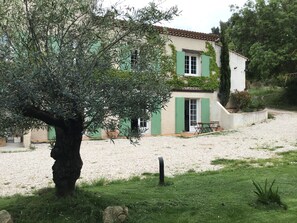 Une maison à deux étages avec des volets verts, une allée en gravier et un grand arbre au premier plan.