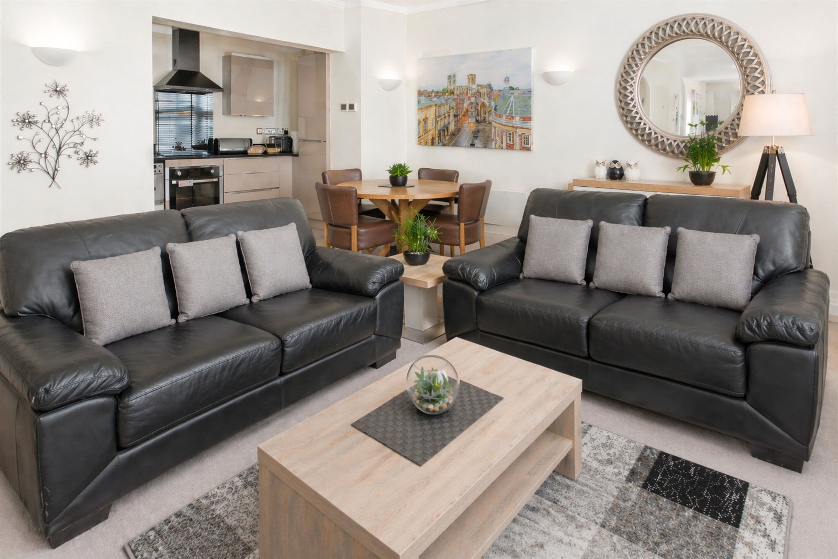 A modern living room with two black leather sofas, a wooden coffee table, a dining table with chairs, a kitchen in the background, and a large mirror on the wall.