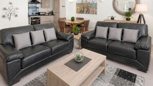 Un salon moderne avec deux canapés en cuir noir, une table basse en bois, une table à manger avec des chaises, une cuisine en arrière-plan et un grand miroir au mur.