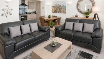 A modern living room with two black leather sofas, a wooden coffee table, a dining table with chairs, a kitchen in the background, and a large mirror on the wall.