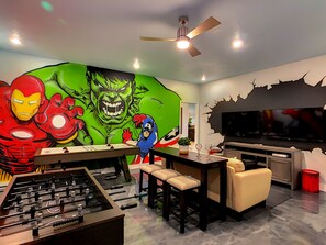 Una habitación con una mesa de fútbol de mesa, un televisor grande y un mural con superhéroes.