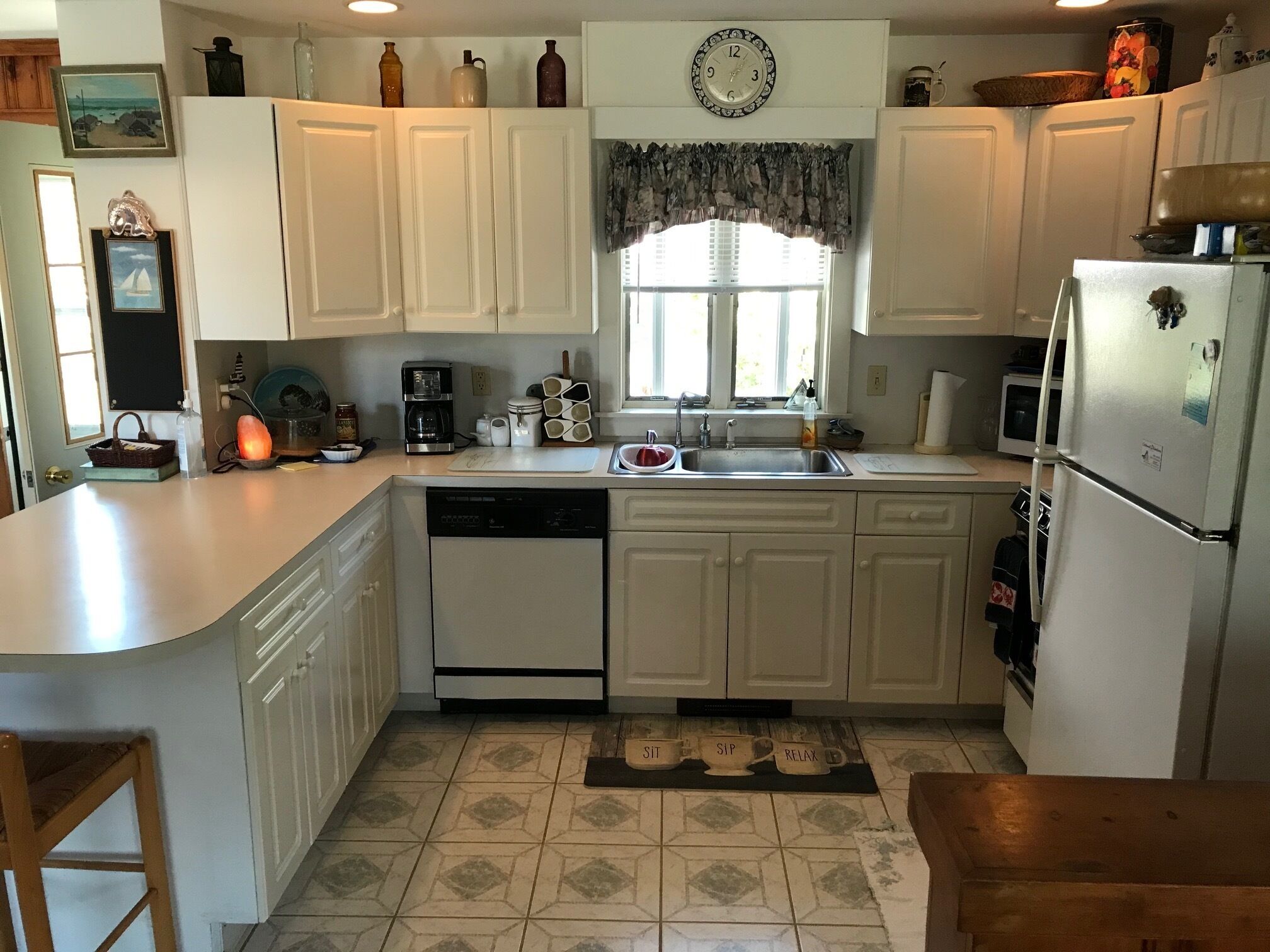 A kitchen with white cabinets, a dishwasher, a refrigerator, a coffee maker, and a window with curtains.