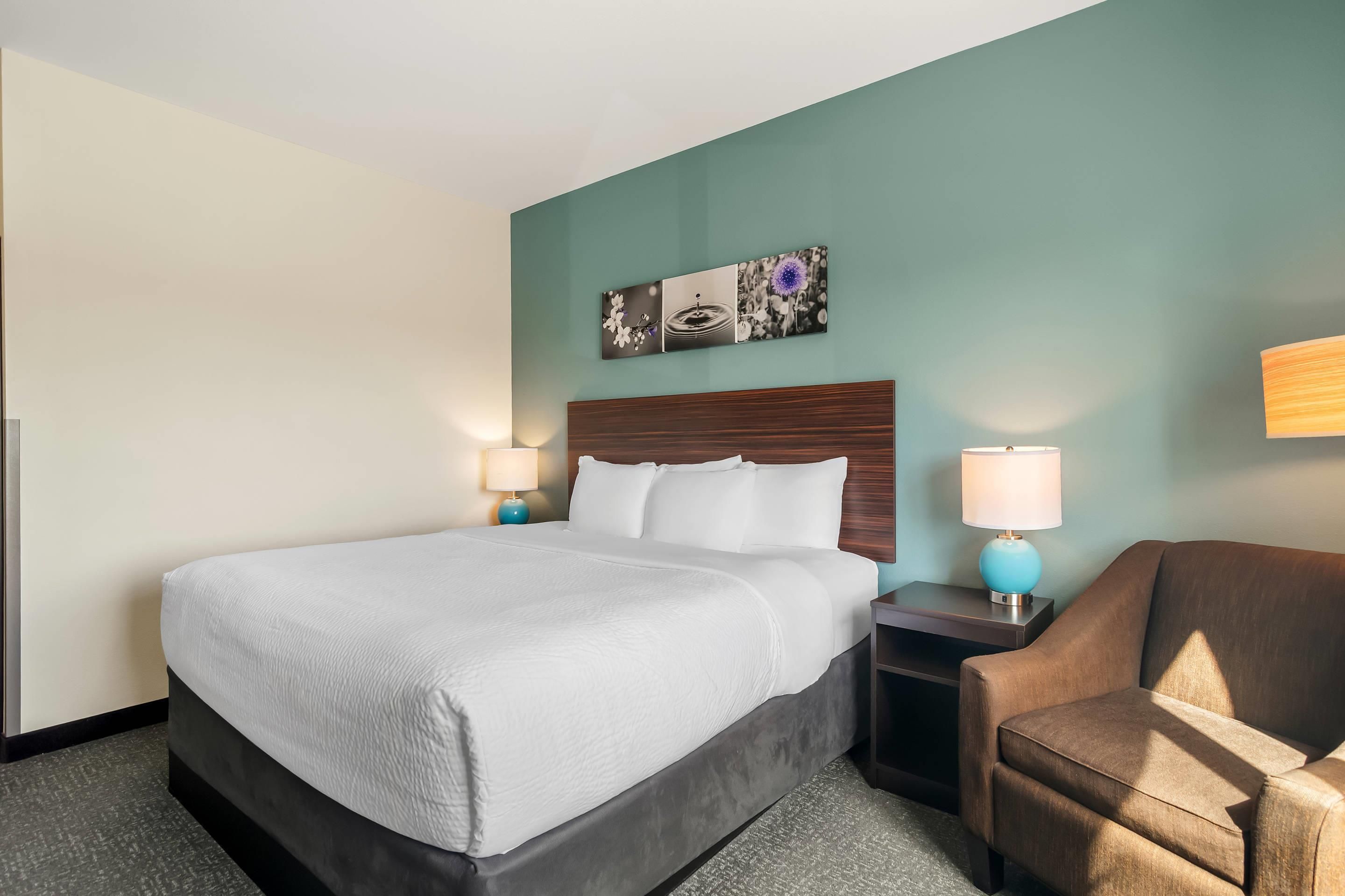 A hotel room with a large bed, two bedside tables with lamps, a brown armchair, and a wall-mounted picture.