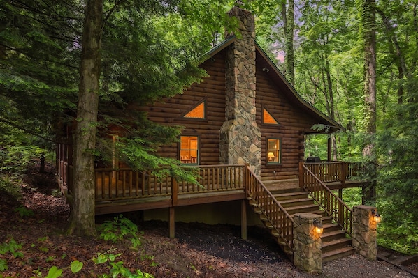 Une cabane en rondins avec une cheminée en pierre, une terrasse en bois et un escalier menant à l’entrée.