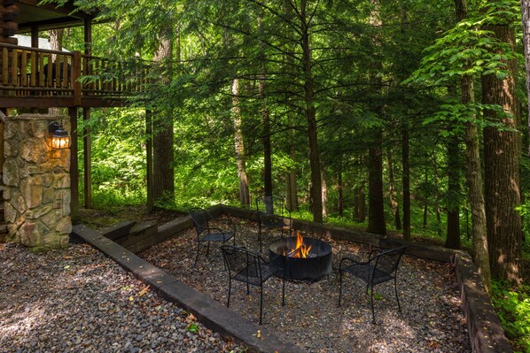 A fire pit with chairs, surrounded by a stone wall and a wooden deck, set in a forested area.
