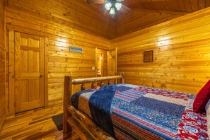 A log cabin room with a wooden floor, a bed with a quilt, a wooden bench, a door, and a sign on the wall.