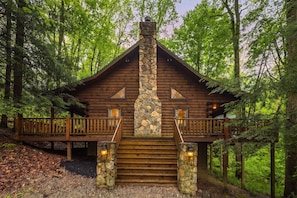 A log cabin with a stone chimney, wooden deck, and a stone staircase leading to the entrance.