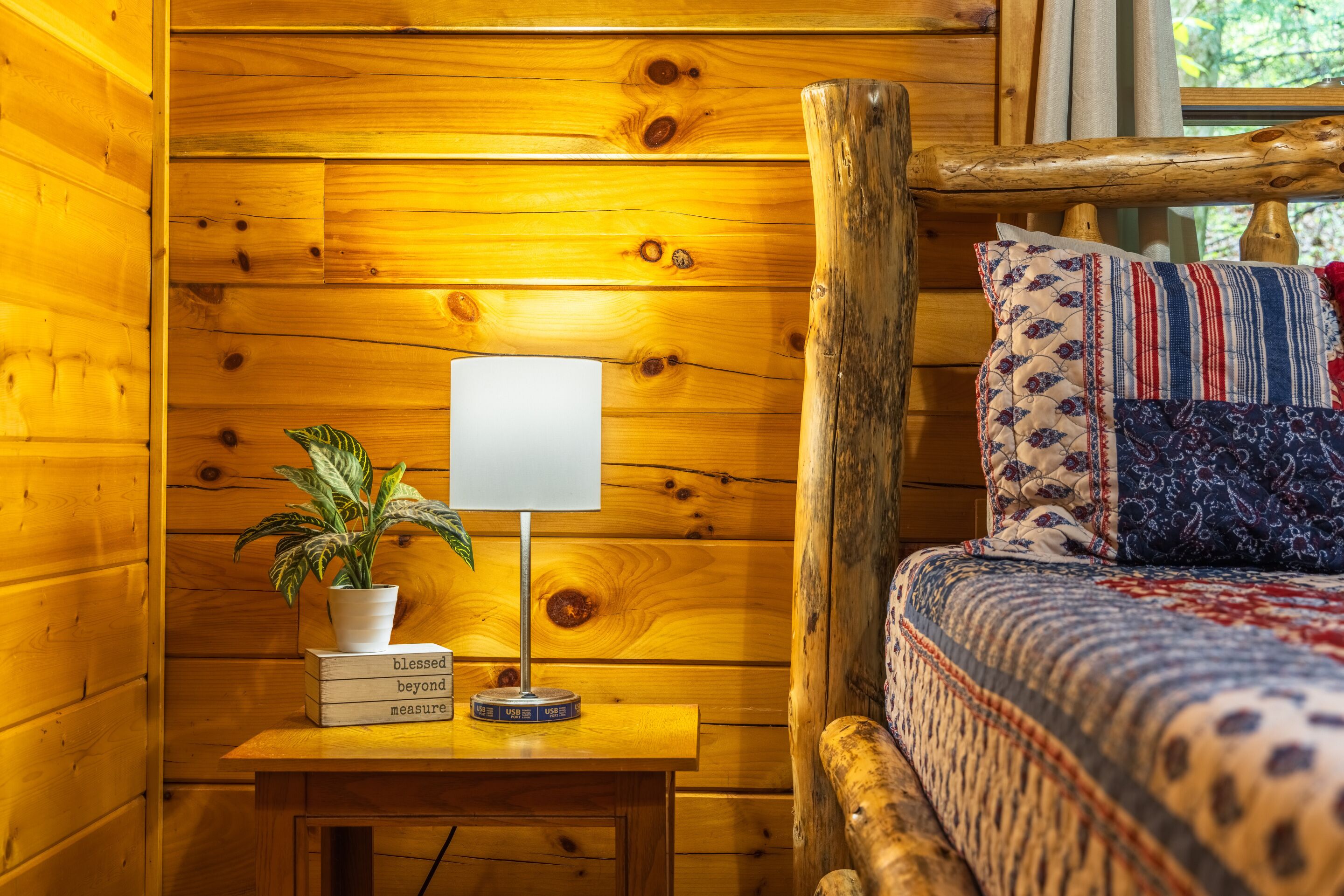 Ein Holzblockhaus-Interieur mit einem Bett, einem Nachttisch mit Lampe, einem Blumentopf und einem Fenster mit Vorhängen.