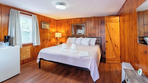Ett rum med träpanel på väggarna, innehållande en säng, ett sängbord, en lampa och en liten köksdel.
