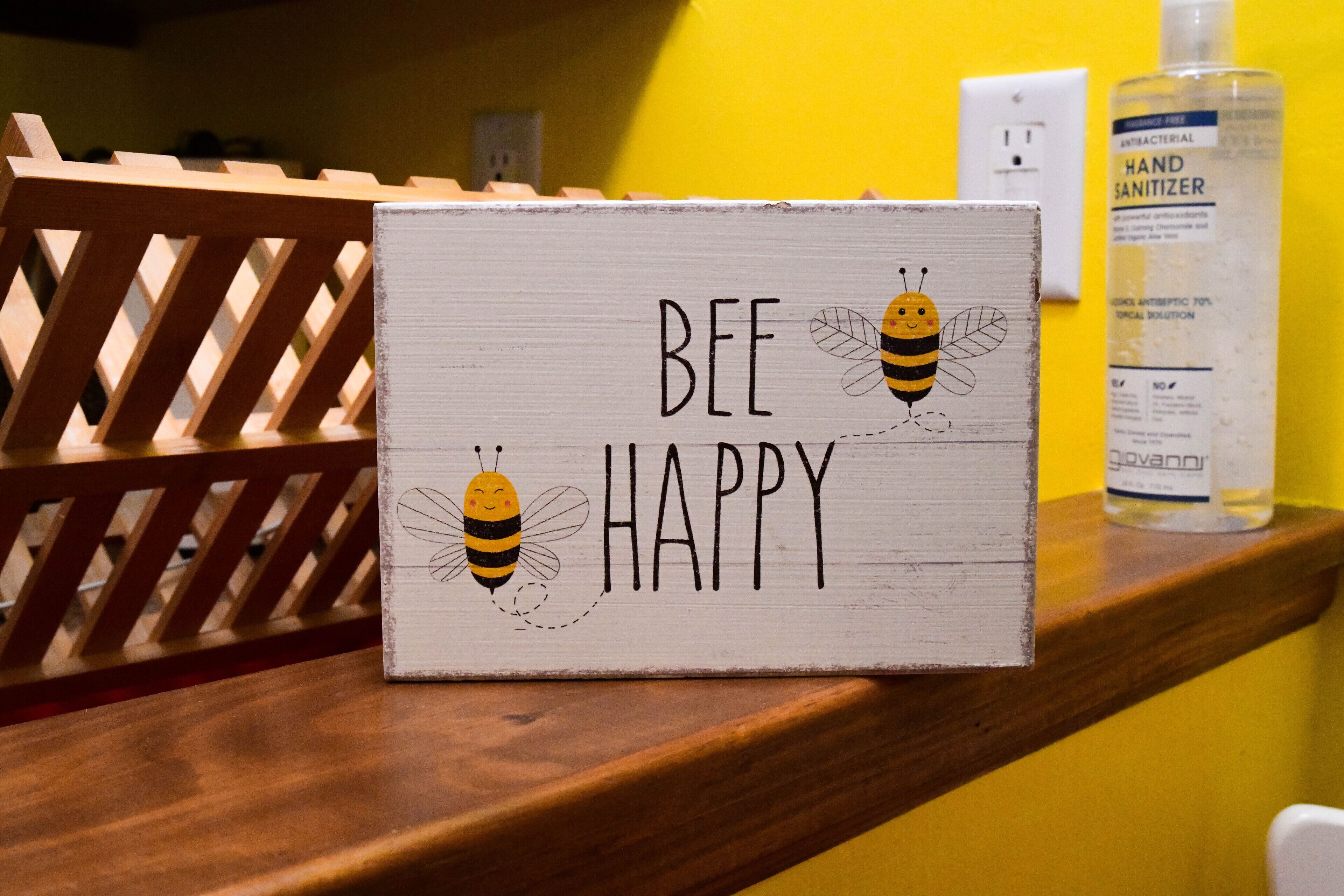 Une enseigne en bois représentant des abeilles et portant l’inscription « SOYEZ HEUREUX » est posée sur une étagère.
