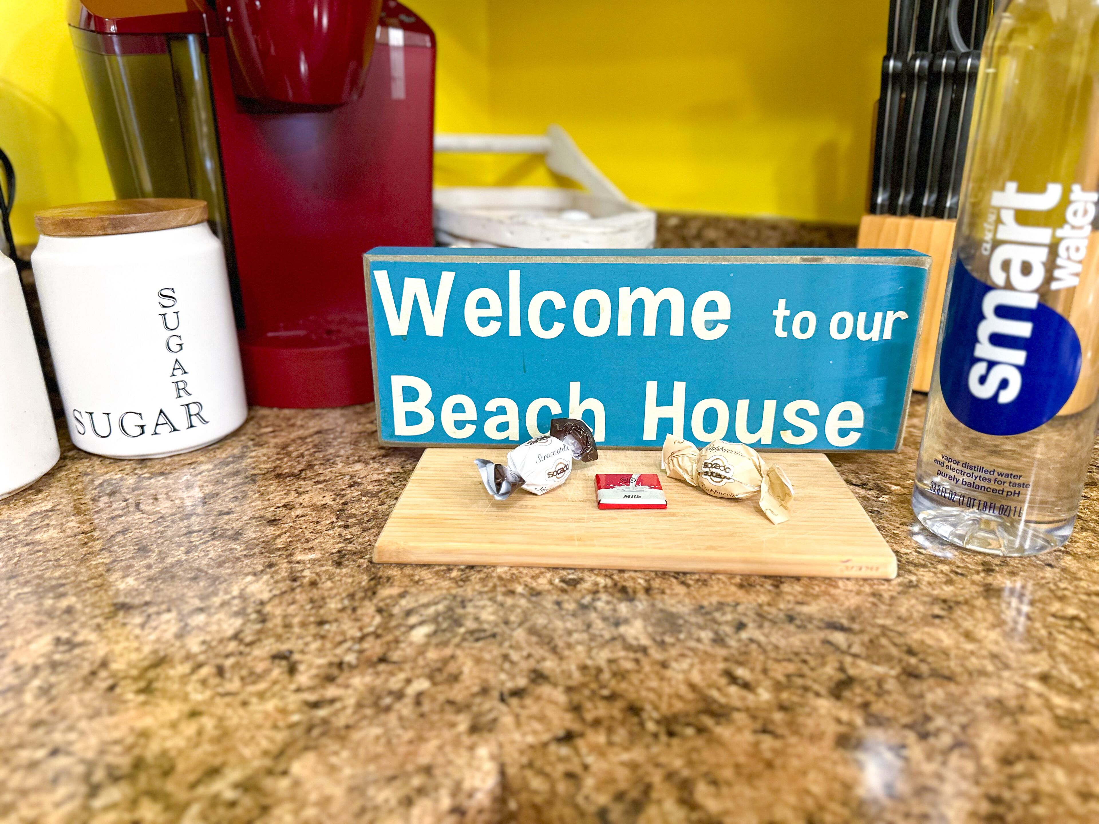 Een keukentafel met een bordje 'Welcome to our Beach House', een suikerpot, een rood apparaat, een fles Smart Water en wat ingepakte snoepjes.