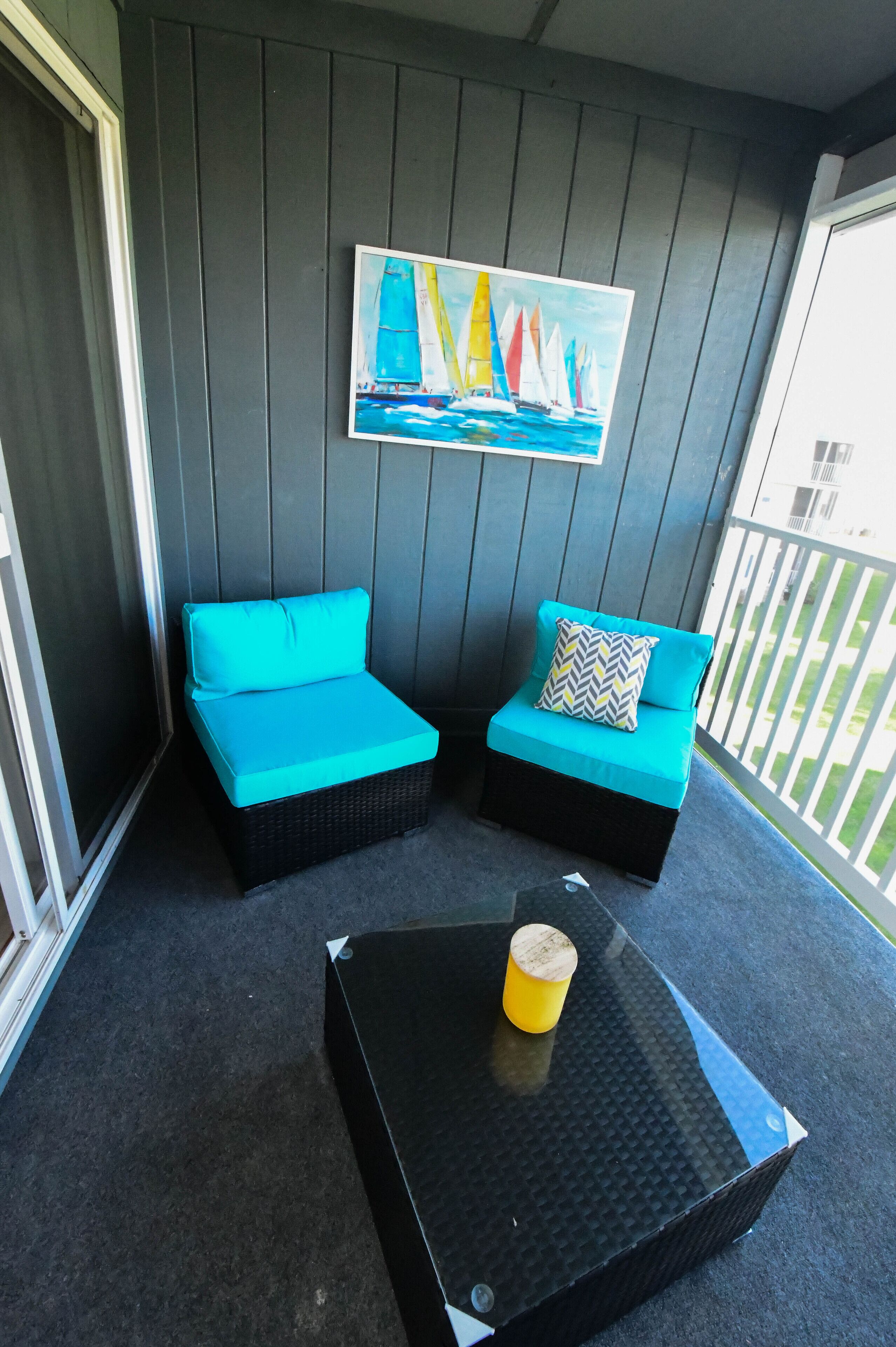 A balcony with two blue cushioned chairs, a black table with a yellow object, and a painting of sailboats on the wall.