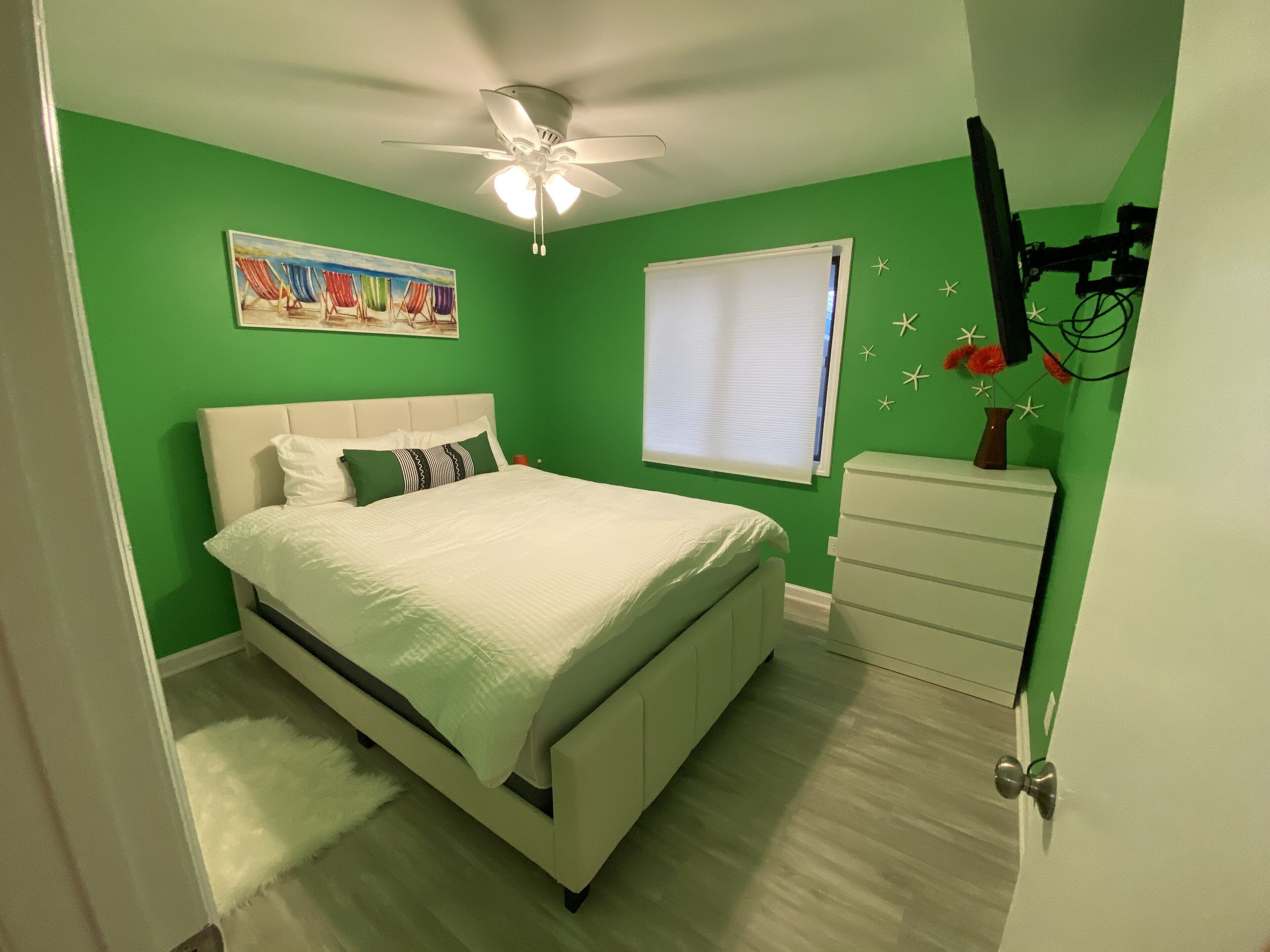 A bedroom with a bed, a ceiling fan, a window with blinds, a dresser, and a painting on the wall.
