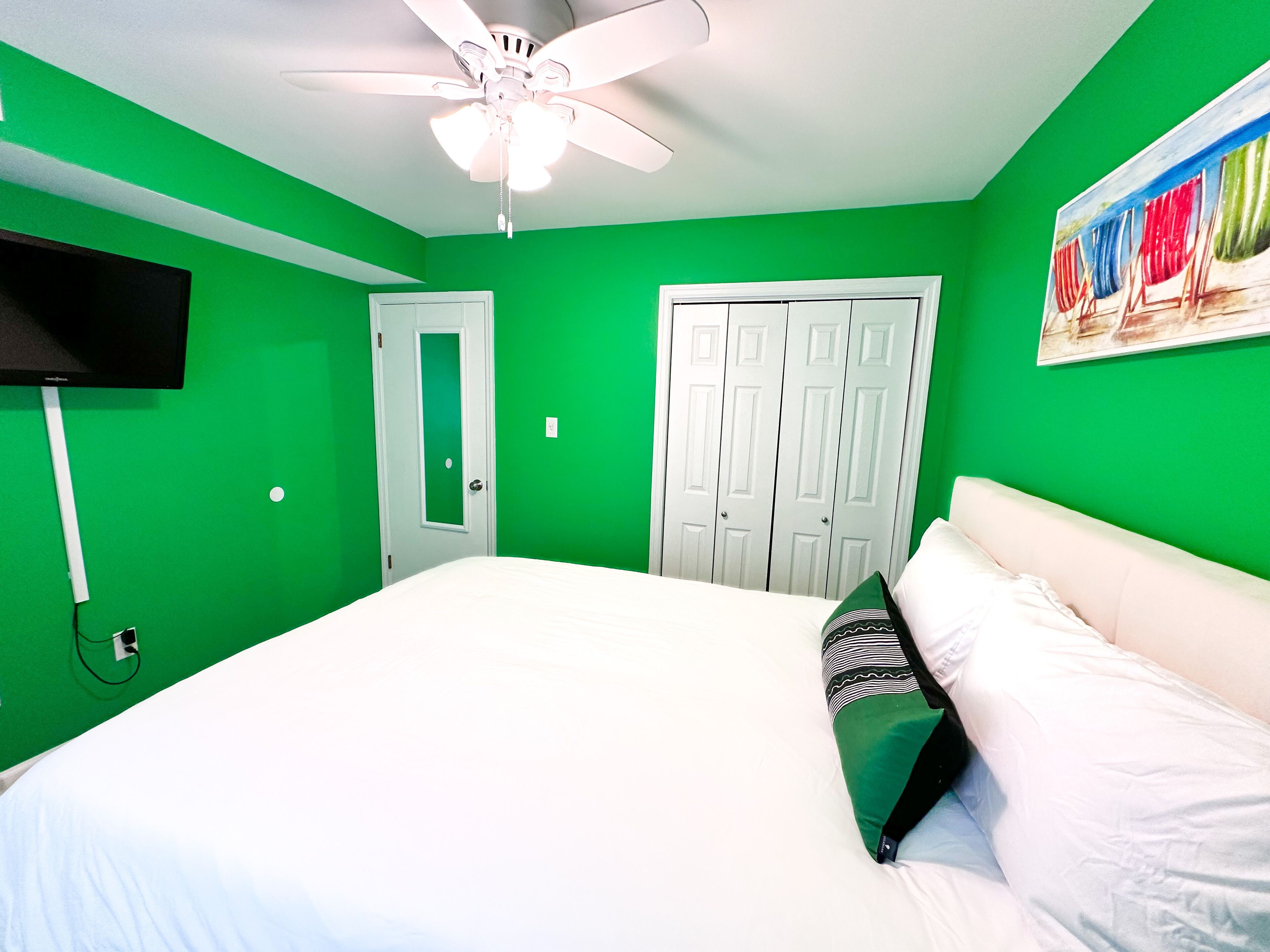 A room with green walls, a ceiling fan, a bed with white bedding, a TV mounted on the wall, and a door with a mirror.