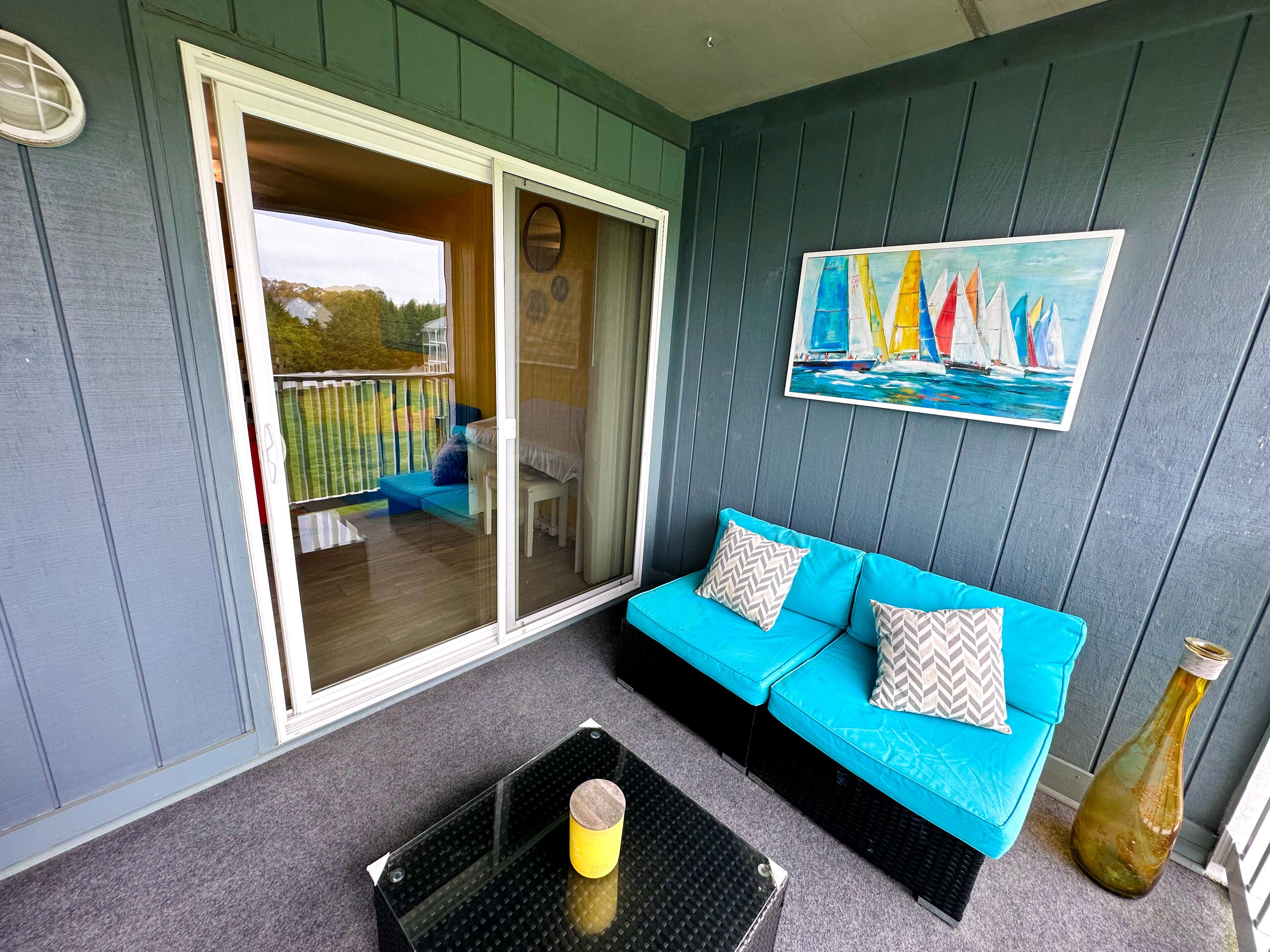 Een kamer met een blauwe bank, een glazen deur naar een balkon, een schilderij van zeilboten en een grote vaas.