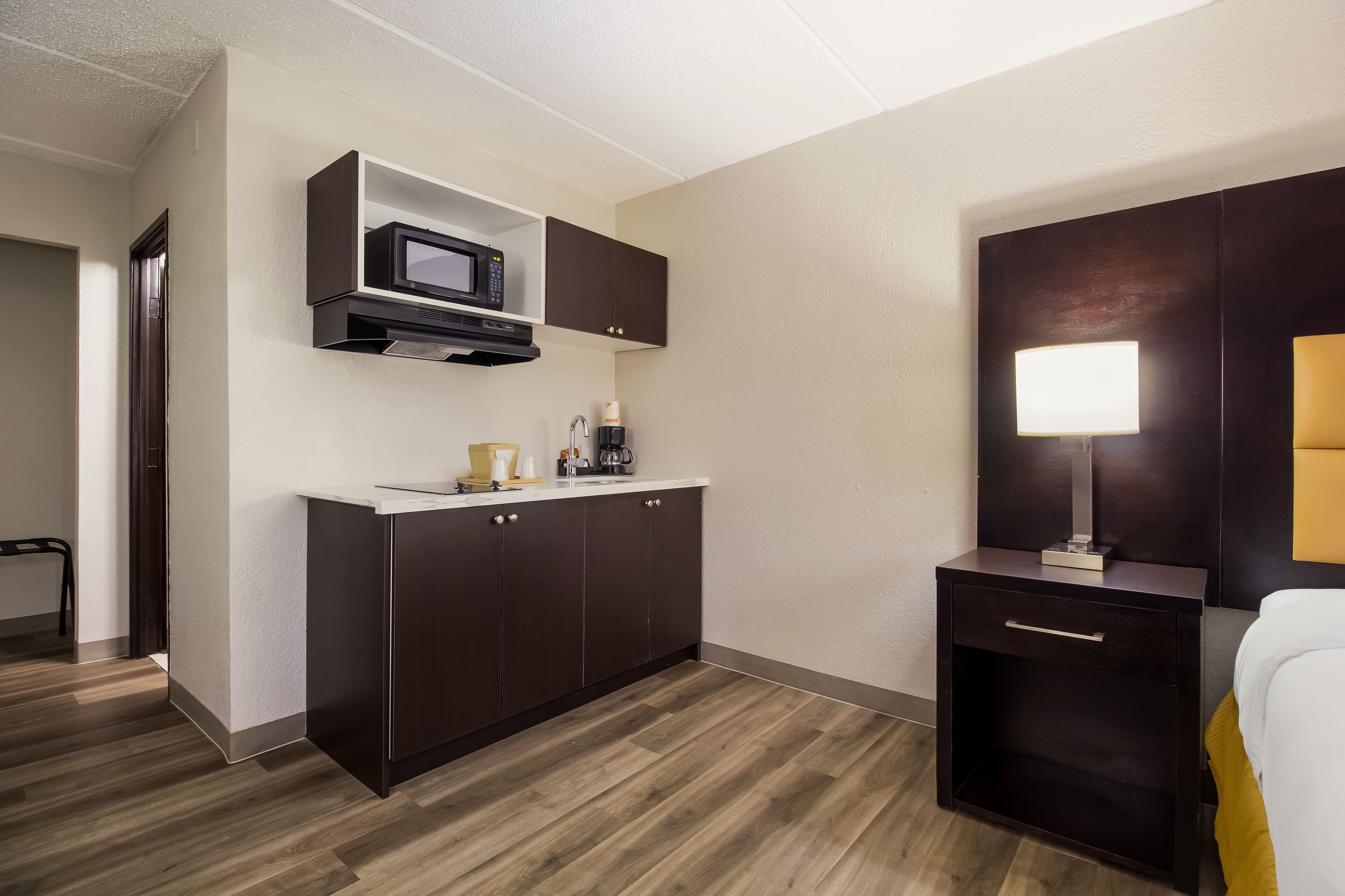 Una habitación de hotel compacta con kitchenette, una mesita de noche con lámpara y una cama con cubrecama blanco.