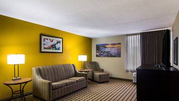 Habitación de hotel con pared amarilla, sofá, silla, mesita con lámpara y televisor.