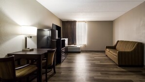 Ein Hotelzimmer mit Holzboden, einer Couch, einem Fernseher, einem Schreibtisch mit Lampe und einem Fenster mit Vorhängen.