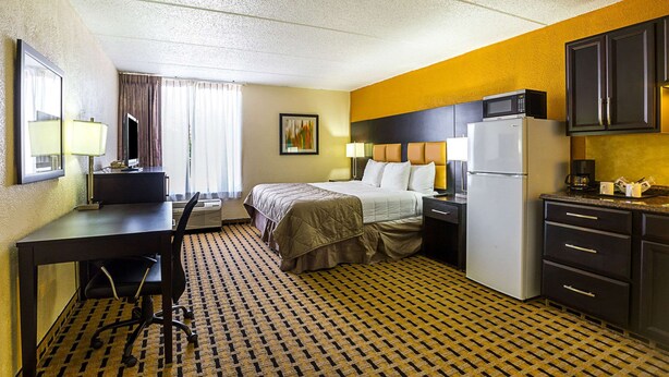 A hotel room with a bed, desk, chair, refrigerator, microwave, and a small kitchen area.