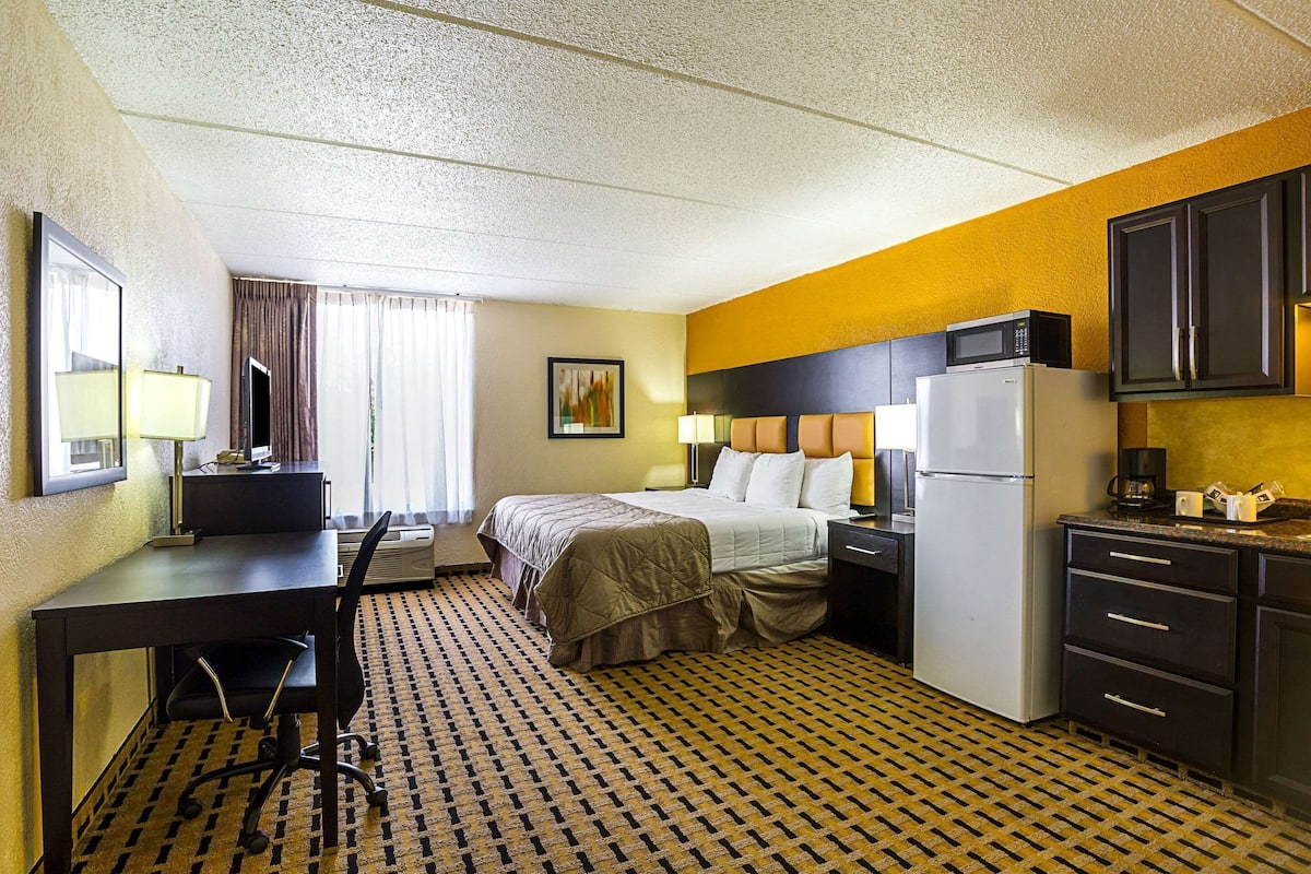 A hotel room with a bed, desk, chair, refrigerator, microwave, and a small kitchenette.