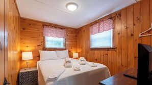 Ett rum med träpanel på väggarna, en säng, två sänglampor, en tv och ett fönster med mönstrade gardiner.