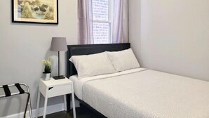 Une chambre à coucher bien rangée, avec un lit simple, une table de chevet avec une lampe, une petite plante en pot et une œuvre d’art encadrée accrochée au mur.