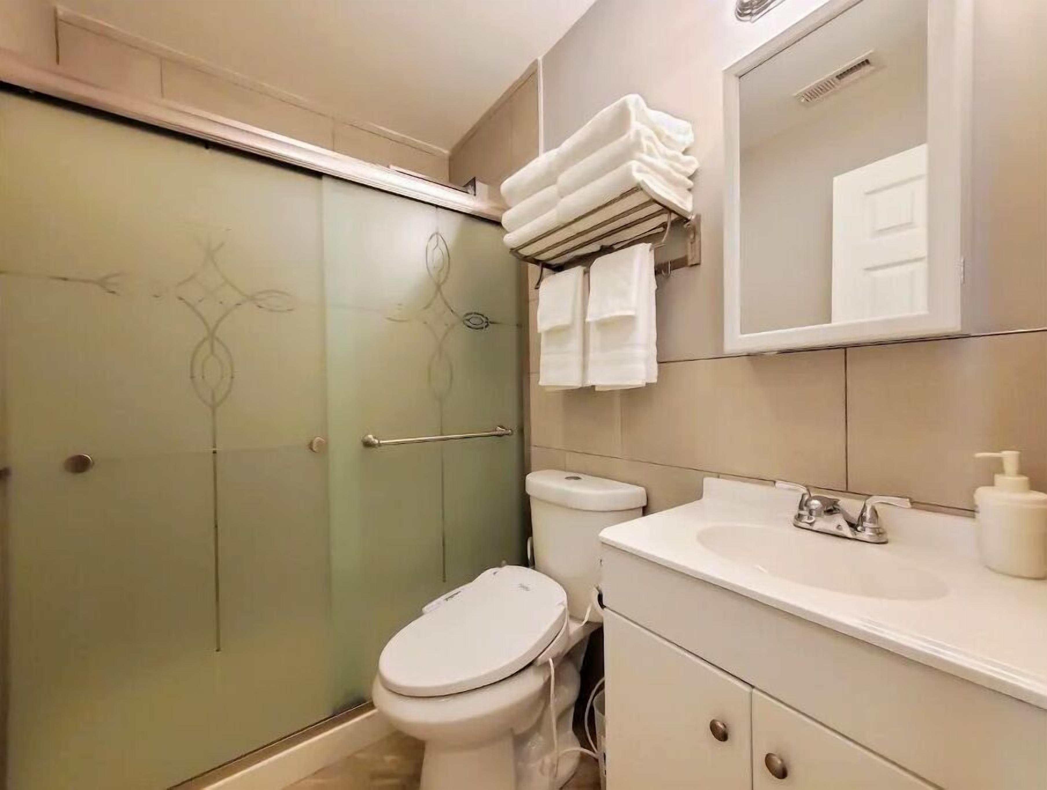 Un baño con mampara de vidrio, inodoro, lavamanos con espejo y toallero con toallas dobladas.