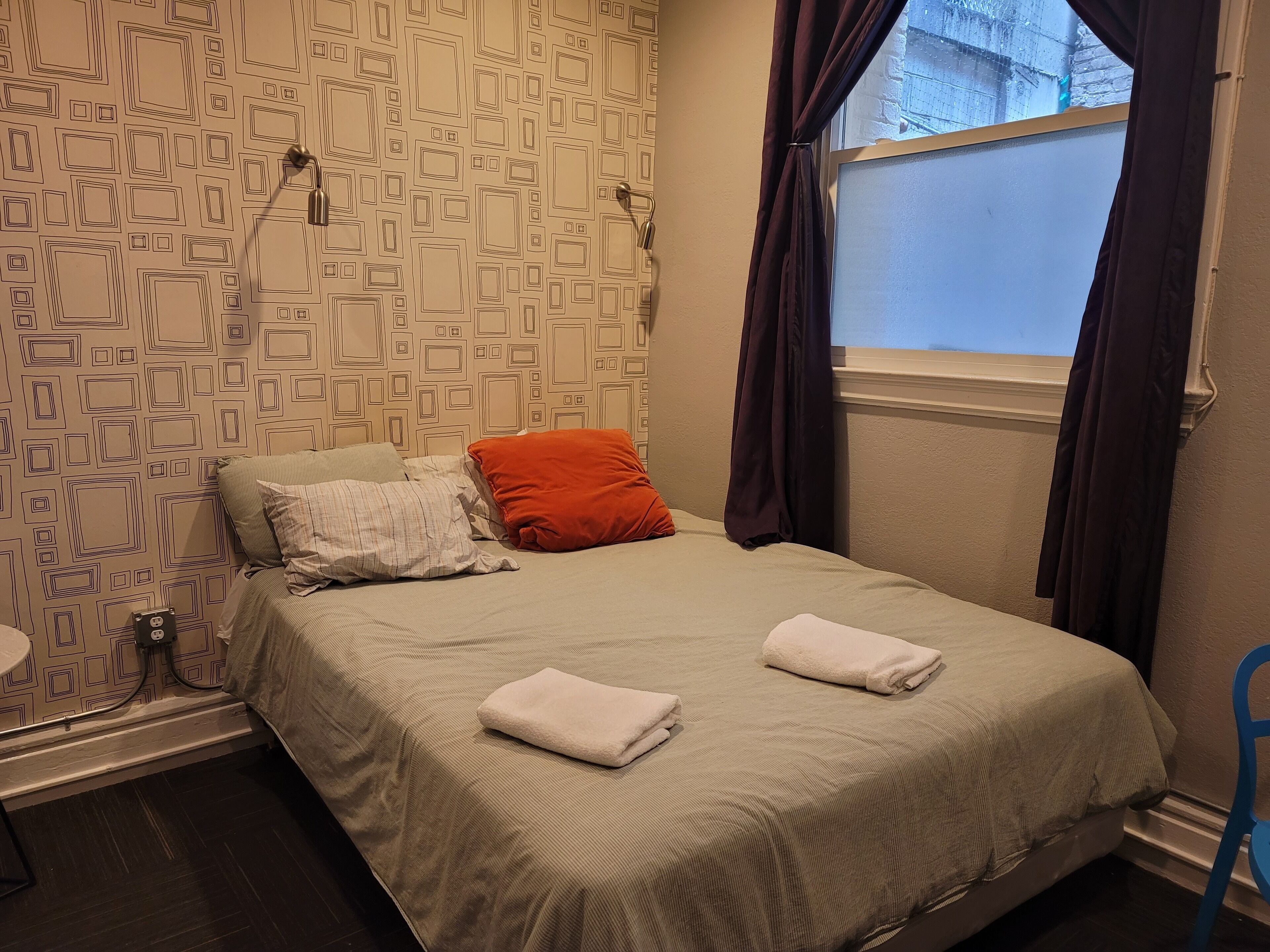 Un lit bien fait, recouvert d’une couvre-lit beige et agrémenté de deux serviettes blanches, un mur à motifs, une fenêtre avec des rideaux foncés et une chaise bleue.