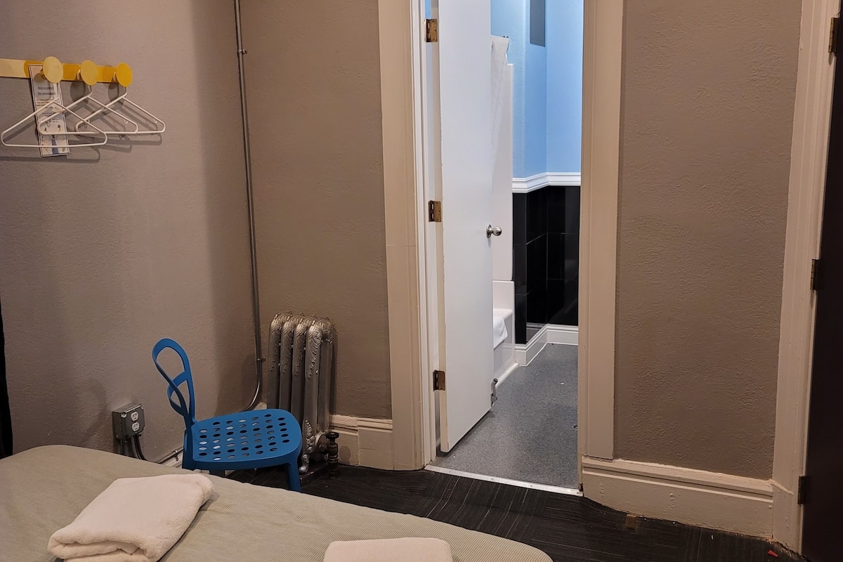 A hotel room with a bed, a blue chair, a radiator, and a door leading to a corridor.
