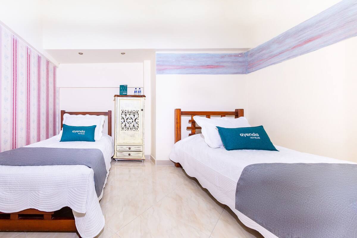Habitación de hotel con dos camas, cada una con un cojín que dice 'ayenda hotels'. Hay un armario y un mueble con objetos encima.