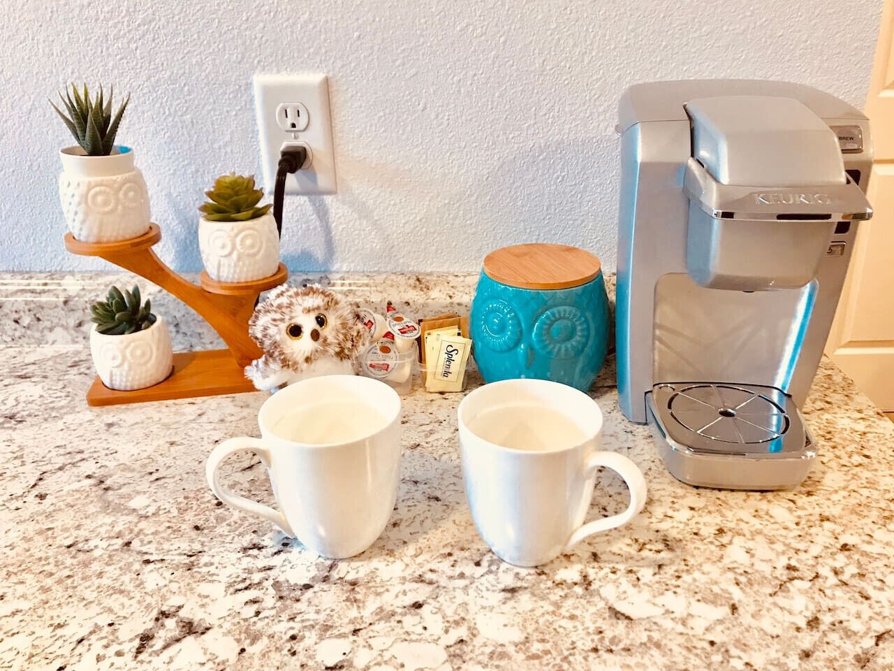 A kitchen counter with two white mugs, a Keurig coffee maker, a small owl figurine, and a blue teapot.