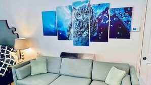 Ein Wohnzimmer mit grauem Sofa, einem gemusterten Sitzkissen und einem an der Wand hängenden Gemälde einer Eule.