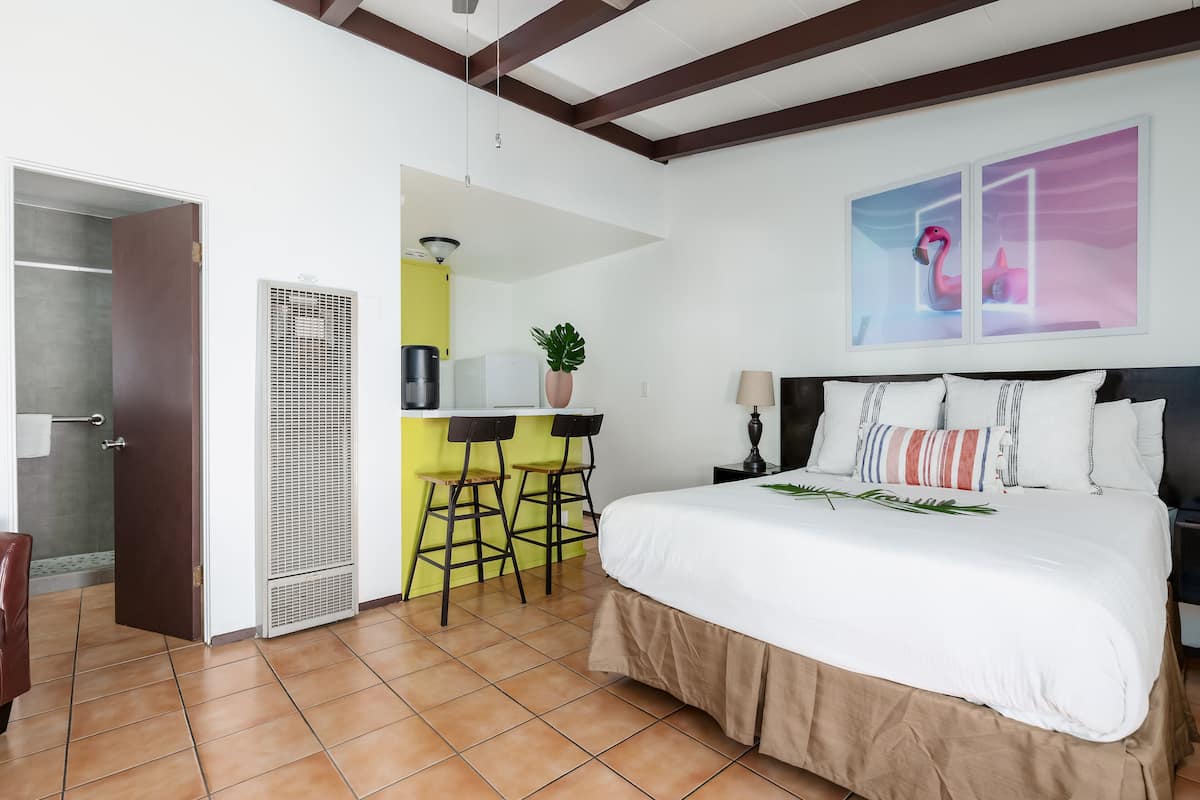 A hotel room with a bed, a desk, two bar stools, a potted plant, and a painting on the wall.