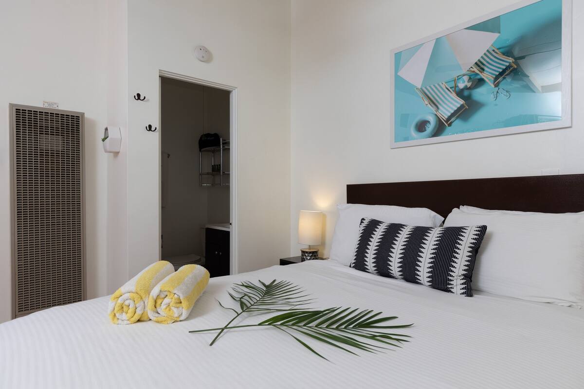A hotel room with a neatly made bed, a wall-mounted air conditioner, a framed artwork above the bed, and a visible door leading to a bathroom.
