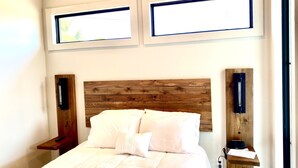 Ein Schlafzimmer mit einem hölzernen Kopfteil, zwei Nachttischen, einem Bett mit weißer Bettwäsche und zwei Fenstern mit Jalousien.