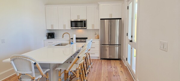 A modern kitchen with white cabinets, a stainless steel refrigerator, a microwave, and a long island with bar stools.