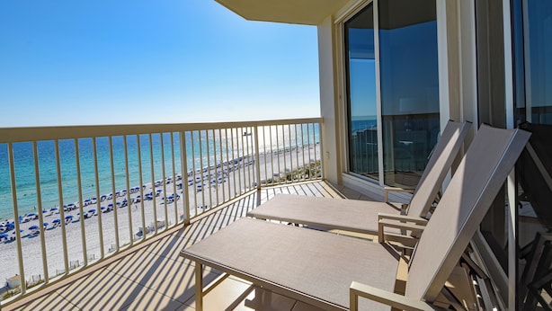 A balcony with a view of the beach and ocean. There are two lounge chairs and a table.