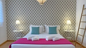 Une chambre d’hôtel avec un papier peint à motifs, deux tables de chevet avec des lampes, un lit avec une literie blanche et des coussins turquoise, et un plaid rose.