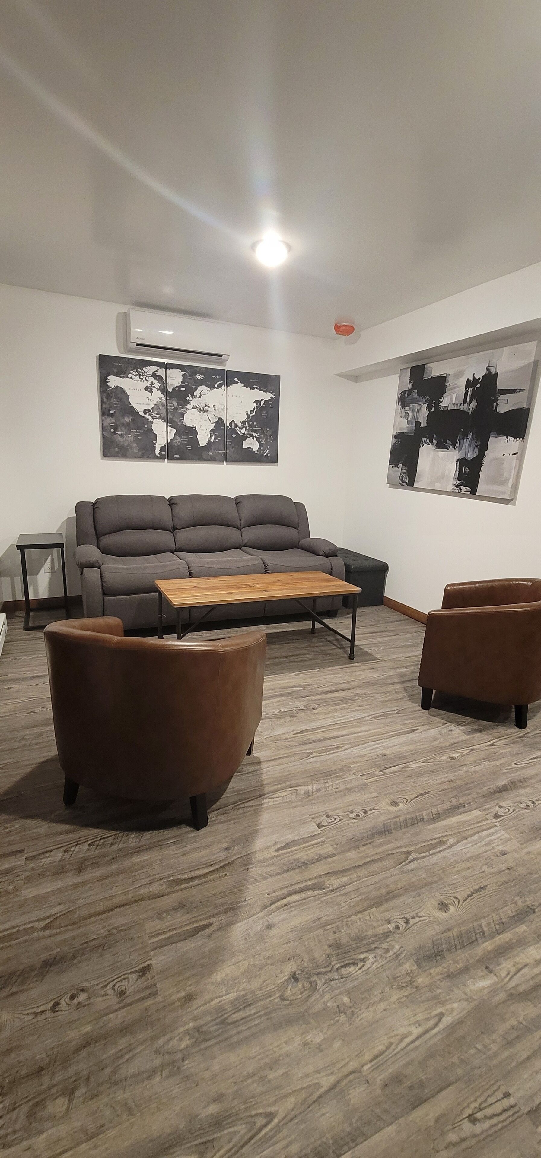 Una sala de estar moderna con un sofá gris, una mesa de centro de madera y dos sillones marrones. Un mapa mundial y un cuadro abstracto decoran las paredes.