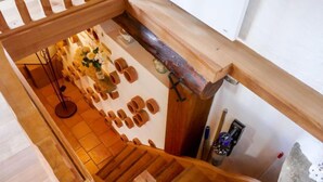 Una escalera de madera que baja a un nivel inferior con piso de baldosas, una pequeña mesa con un jarrón de flores y una pared con varios jarrones de barro.