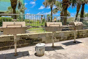 Outdoor dining area with barbecue grills and trash bin, surrounded by palm trees.