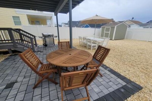 A covered patio with a round wooden table and chairs, a grill, and a shed in the background. - 2-Family Beach House - Ortley Beach, NJ (Seaside Heights)