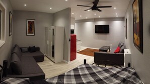 Ein modernes Hotelzimmer mit einer Couch, einem Bett mit kariertem Überwurf, einem Deckenventilator, Einbauleuchten, einem Flachbildfernseher und einem Kühlschrank.