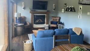 Un salon chaleureux avec une cheminée, des canapés bleus, une table basse en bois et un téléviseur fixé au-dessus de la cheminée.