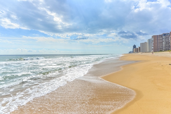 A beach with a sandy shore and ocean waves, with buildings in the background.
