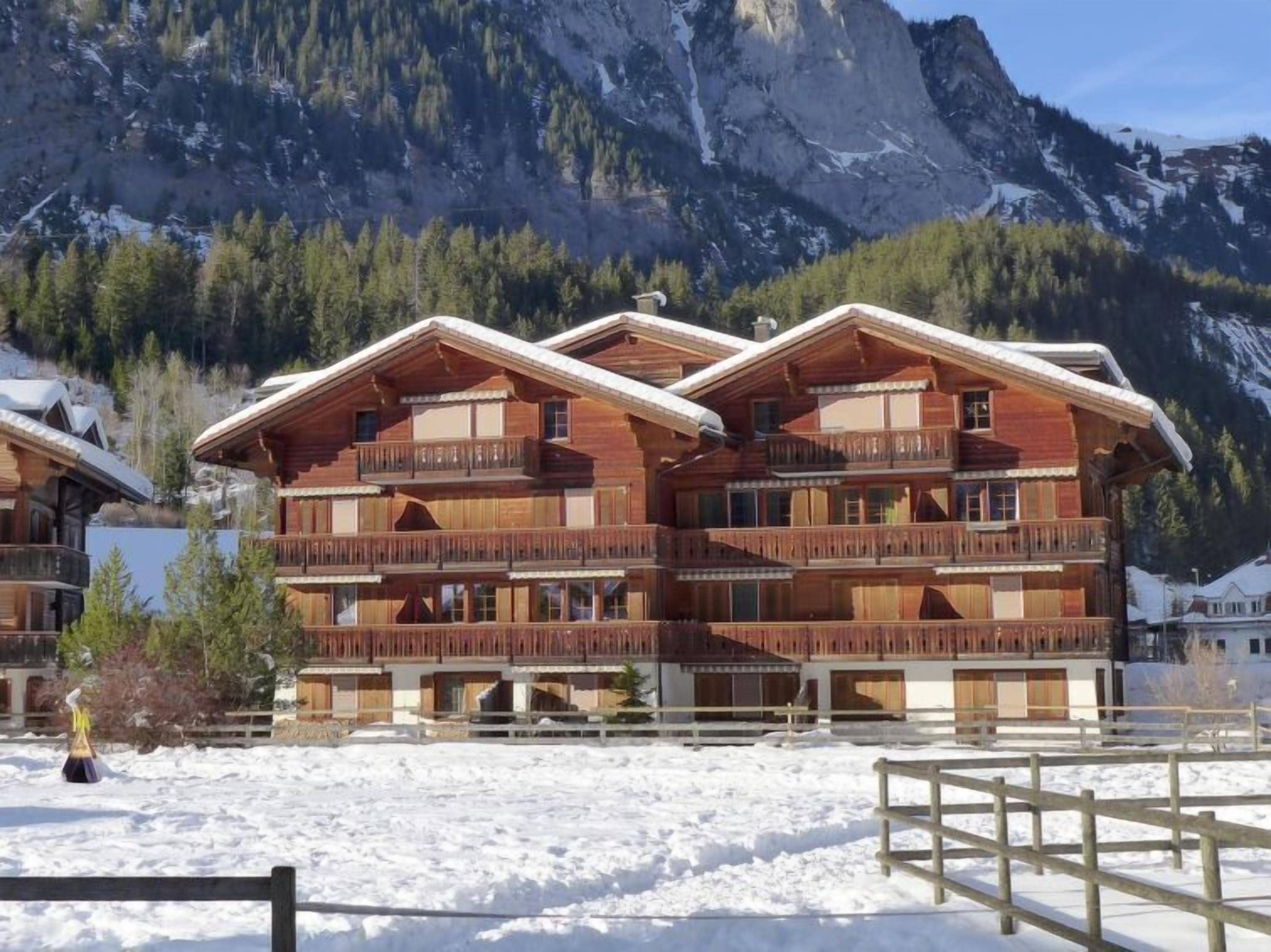Un chalet en bois à plusieurs étages, doté de balcons, qui se dresse sur un fond de paysage montagneux.