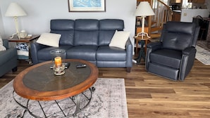Ein Wohnzimmer mit einem blauen Ledersofa, einem Holztisch und einem schwarzen Ledersessel. An der Wand hängt ein gerahmtes Bild, und im Hintergrund ist eine Treppe zu sehen.