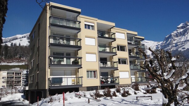 A multi-story building with balconies, a snow-covered ground, and mountains in the background. - Erlenrain Wohnung 722E Appartement/Fewo 2-Bettwohnung by Interhome (Engelberg)